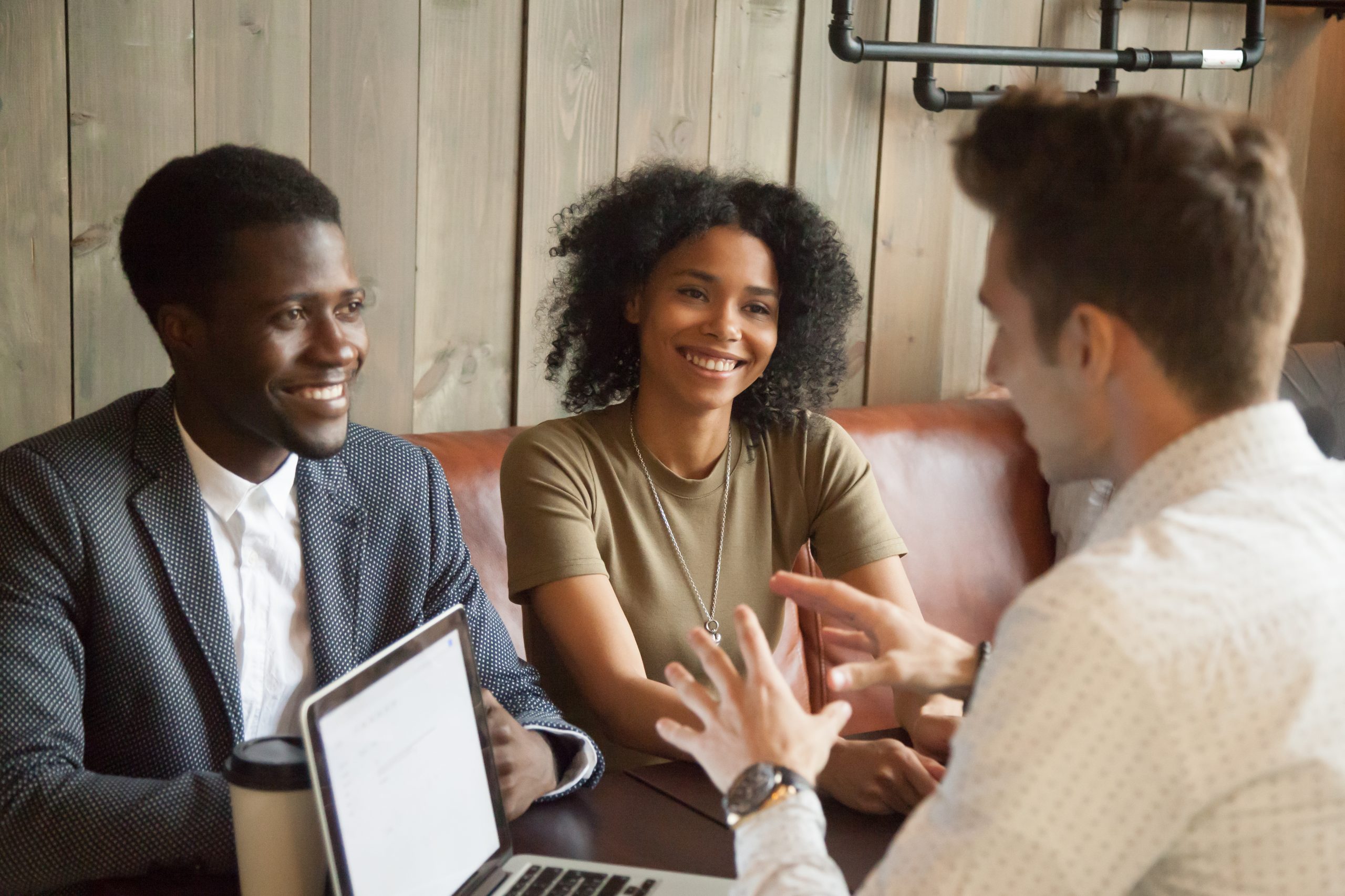 Caucasian architect talking to smiling African American couple, explaining housing project idea in coffee shop, male realtor consulting black clients on property purchase, having conversation in cafe